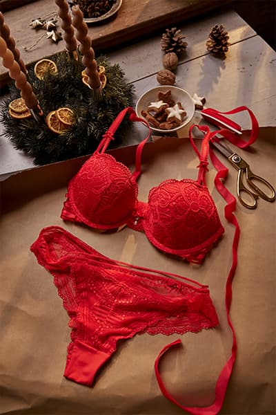 Weihnachtliche Dekoration und rote Spitzenunterwäsche auf einem Tisch, neben einer Schere und rotem Geschenkband.
