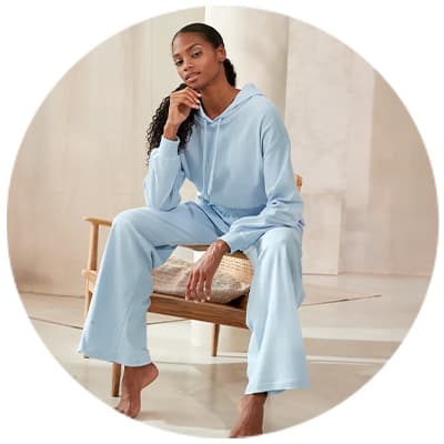 Frau in hellblauem Hoodie und Jogginghose sitzt barfuß auf einem Stuhl in einem minimalistischen Raum.