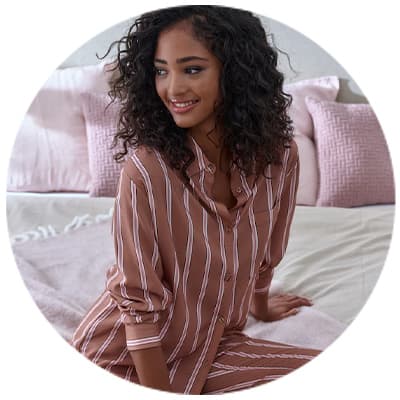 Frau mit lockigem Haar sitzt auf Bett in gestreifter Pyjamahose und passendem Oberteil, rosa Kissen im Hintergrund