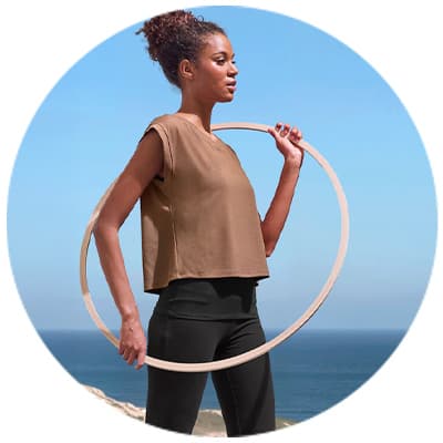 Frau in braunem Oberteil und schwarzen Leggings hält einen Hula-Hoop-Reifen vor einem Meereshintergrund