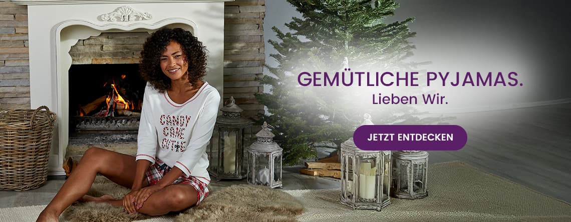 GEMÜTLICHE PYJAMAS. Lieben Wir. Jetzt entdecken – Frau in Pyjama sitzt vor Kamin und Weihnachtsbaum.