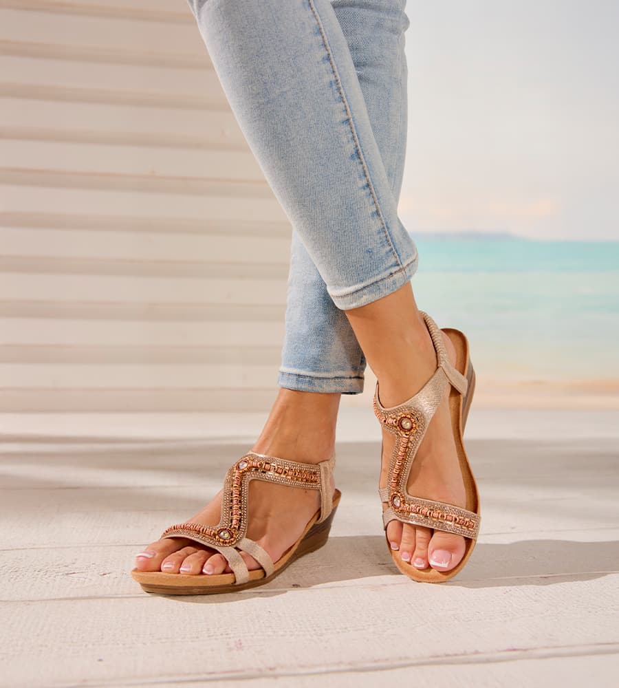 Füße in Sandalen mit Glitzersteinen, Frau trägt hellblaue Jeans, Hintergrund mit Strand und Meer.