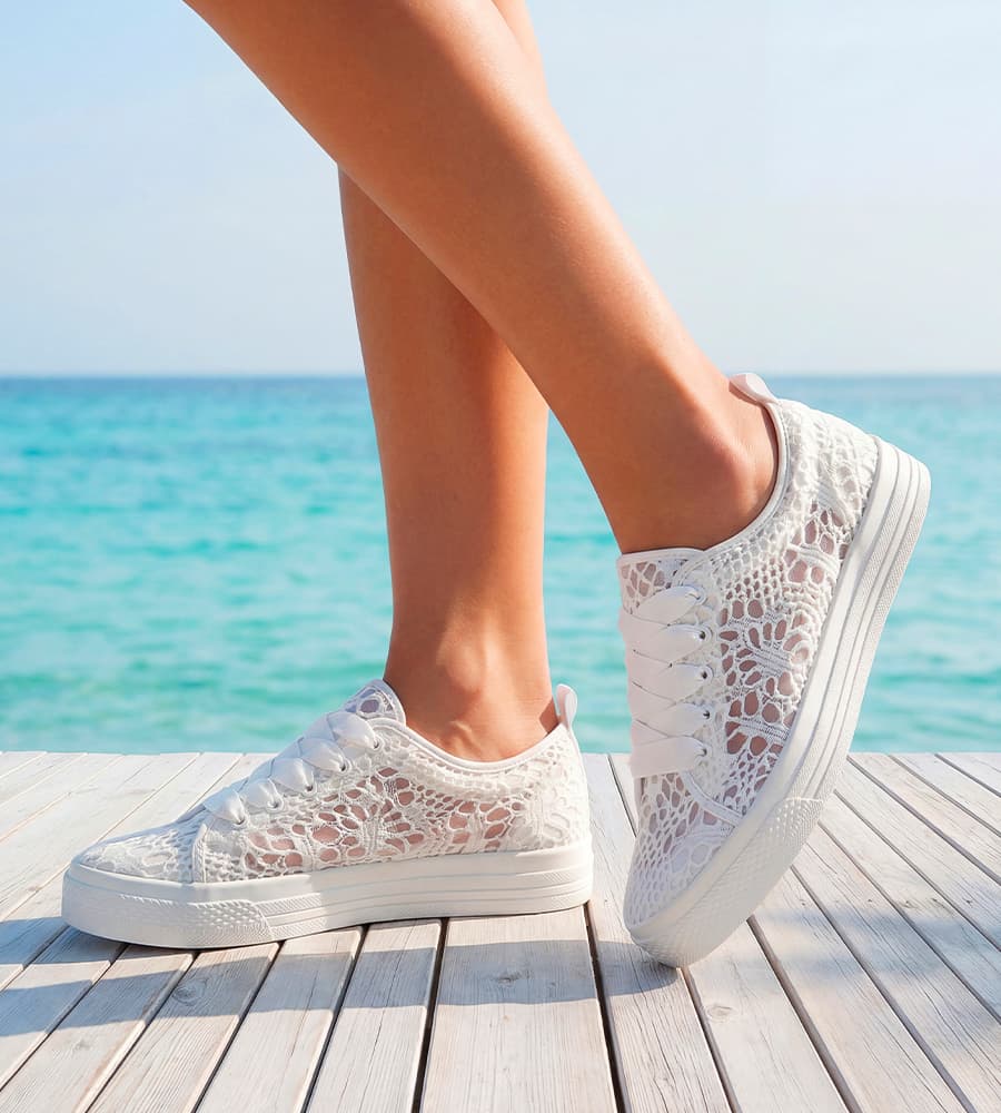 Füße in weißen Sneakern mit Spitzenmuster stehen auf Holzsteg vor türkisblauem Meer