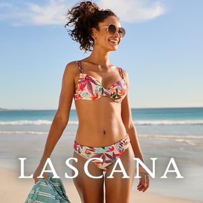 LASCANA – lachende Frau im gemusterten Bikini geht am Strand entlang und hält ein Handtuch in der Hand