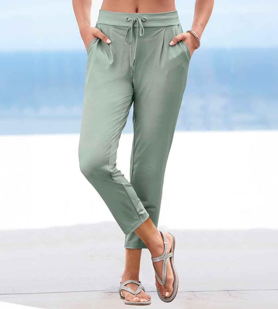 Frau trägt mintgrüne 7/8-Hose mit Bindeband und Eingrifftaschen, dazu Sandalen, steht vor unscharfem Meereshintergrund